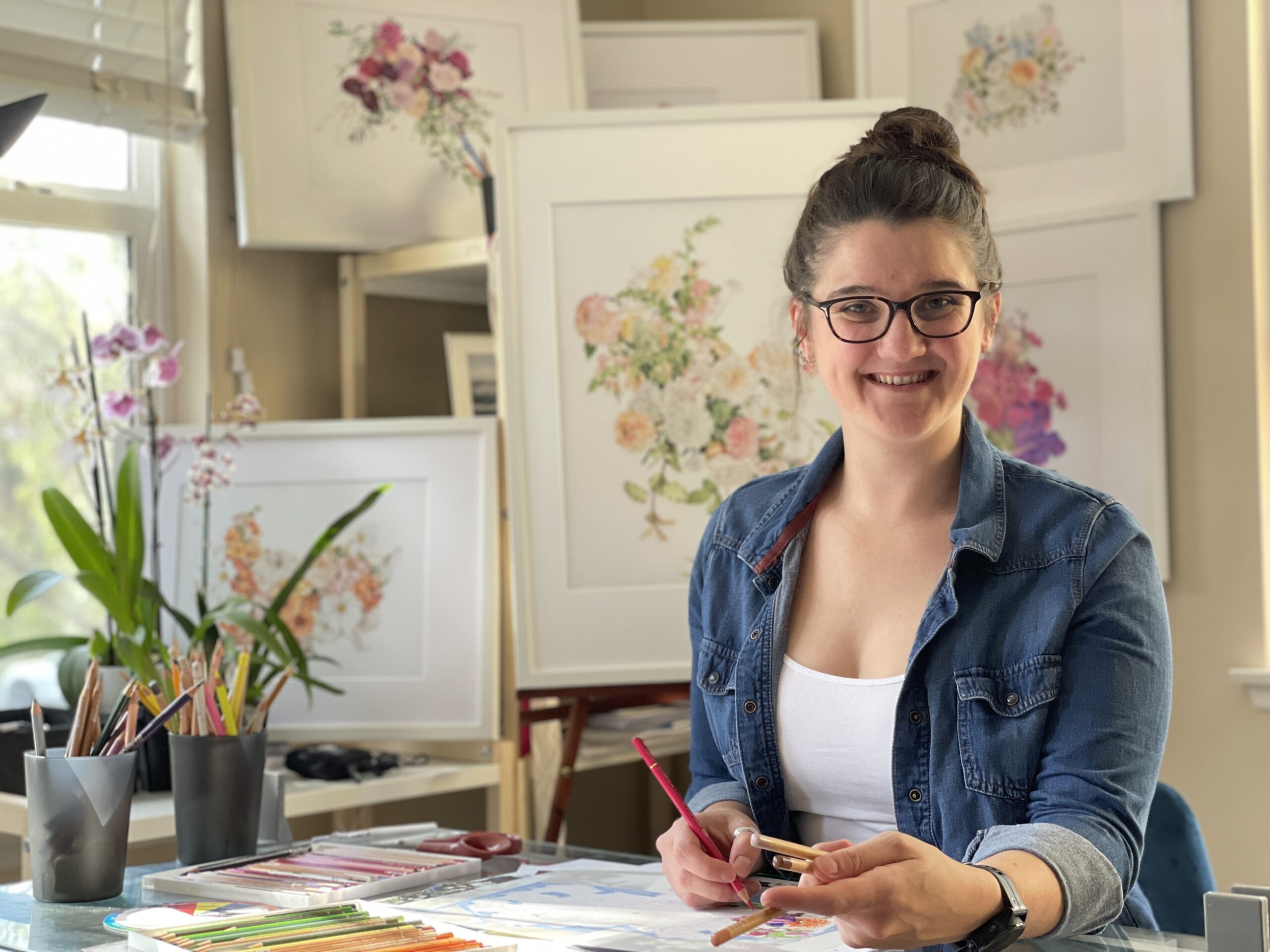 Picture of women sitting at a desk with color pencils in front of pictures she has drawn